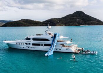 Large white luxury yacht anchored in clear blue water near an island, featuring an inflatable slide and people swimming and paddleboarding nearby—an unforgettable maritime experience awaits.