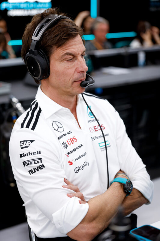A man in a Mercedes-AMG Petronas F1 team shirt, wearing an IWC headset, sits with arms crossed in a Formula 1 garage environment.