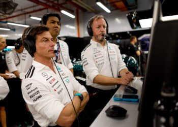Three men in Mercedes-AMG Petronas team uniforms wearing headsets watch monitors intently inside a Formula 1 garage, with Toto Wolff focused on strategy alongside his team.