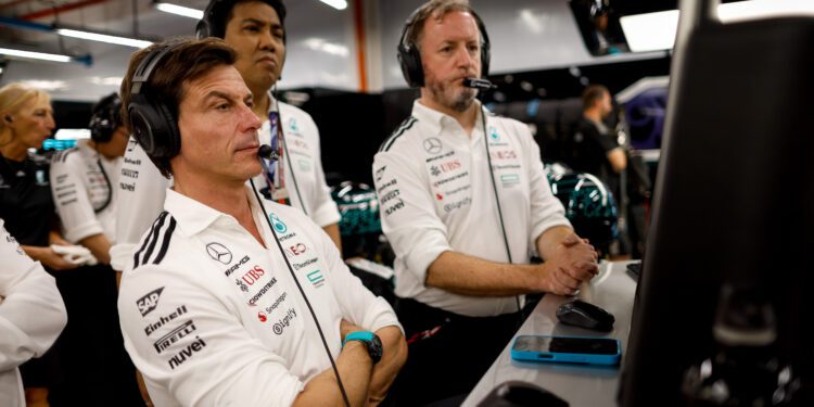 Three men in Mercedes-AMG Petronas team uniforms wearing headsets watch monitors intently inside a Formula 1 garage, with Toto Wolff focused on strategy alongside his team.