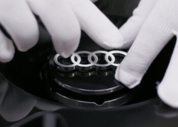 A person wearing white gloves holds a car part featuring the silver Audi logo with four interlinked rings, as seen in a making-of video for a concept car.