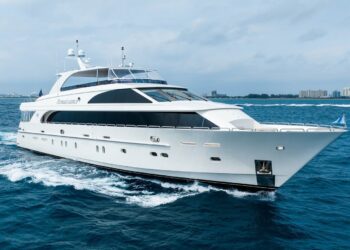 A large white luxury yacht cruises through blue ocean waters near a distant coastline with buildings under a cloudy sky, offering the ultimate maritime experience.