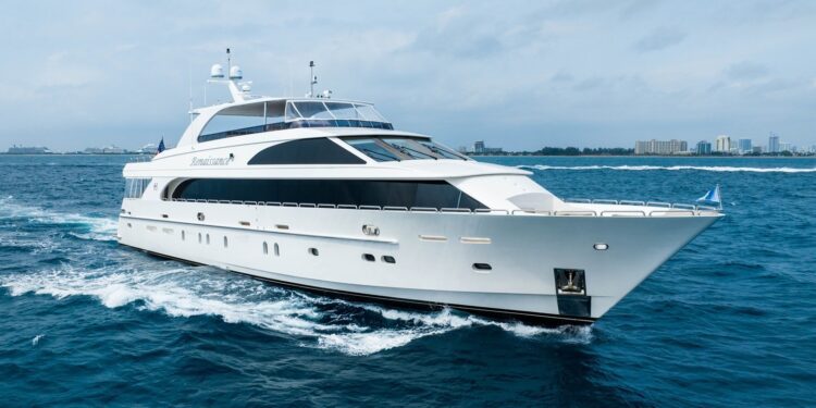 A large white luxury yacht cruises through blue ocean waters near a distant coastline with buildings under a cloudy sky, offering the ultimate maritime experience.