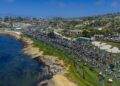 A large crowd gathers on a grassy area by the ocean for the Pebble Beach Concours d’Elegance, celebrating 75 years of automotive artistry, with classic cars on display and buildings in the background under a blue sky.