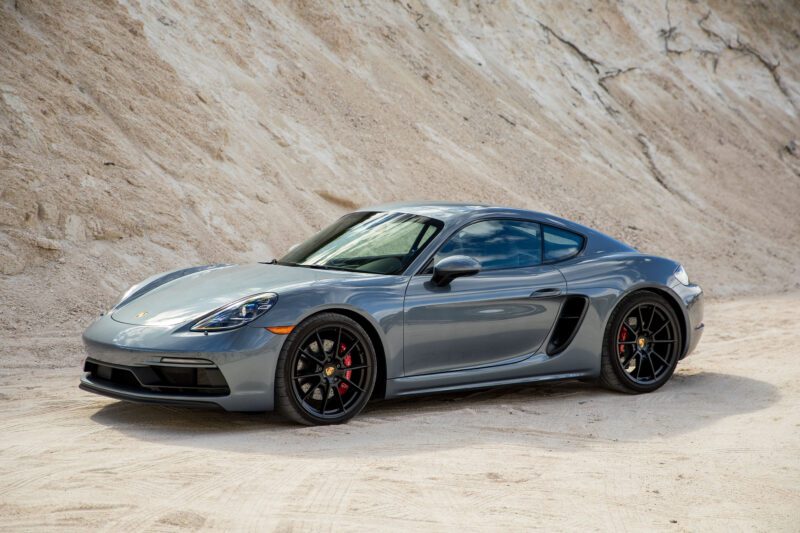 A grey sports coupe with black wheels, reminiscent of the Review: 2025 Porsche 718 Cayman GTS 4.0 Is Close To Flawless, is parked on a sandy surface in front of a rocky, light-colored hill.