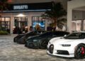 Three Bugatti hypercars are parked in front of Bugatti Broward at night, with people standing nearby and palm trees in the background.