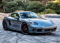 A gray Porsche sports car is parked on a paved area with trees and rocks in the background, echoing the sleek style highlighted in the Review: 2025 Porsche 718 Cayman GTS 4.0 Is Close To Flawless.