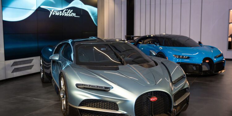 Two Bugatti hypercars, one silver and one blue, are displayed in a modern Bugatti Broward showroom with a digital screen reading "Tourbillon" in the background.