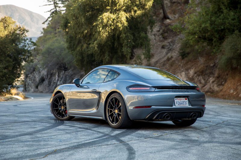A gray Porsche sports car, featured in the review: 2025 Porsche 718 Cayman GTS 4.0 Is Close To Flawless, is parked on a winding mountain road with trees and rocky hillsides in the background.
