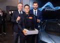 Three men in suits stand together at an indoor event; the man in front holds a Bugatti-branded trophy, with a hypercar and a "Tourbillon" sign in the background.
