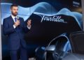 A man in a blue suit speaks into a microphone next to a Bugatti hypercar and a large screen displaying the word "Tourbillon.