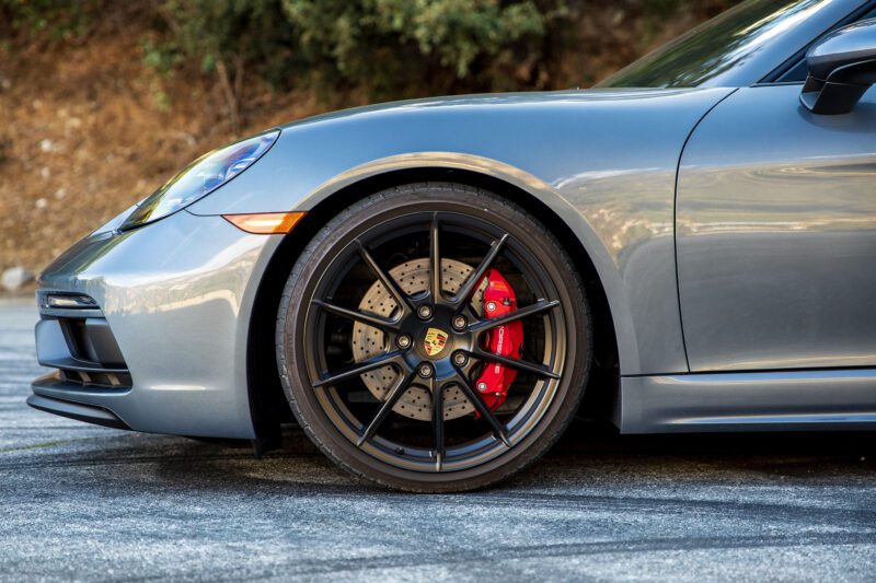 Close-up of the front left wheel of a silver Porsche sports car, as seen in our Review: 2025 Porsche 718 Cayman GTS 4.0 Is Close To Flawless, highlighting the black alloy wheel, red brake caliper, and drilled brake disc.