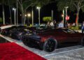 Three luxury sports cars, including a stunning Bugatti hypercar, are parked on a paved lot at night near a red carpet, with people gathered and trees in the background.