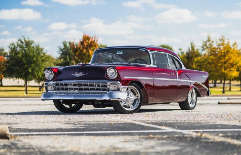 A classic maroon and black vintage car, a true gem among classic muscle cars, is parked in an empty lot with trees and a blue sky in the background.