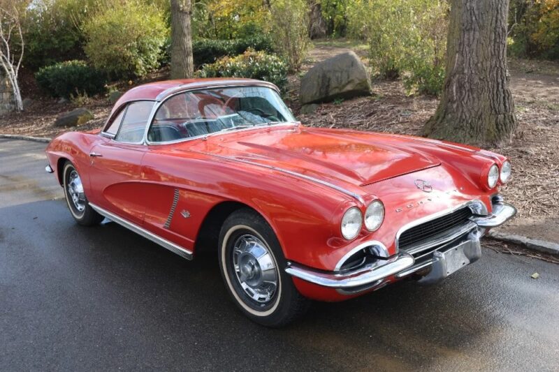 A red classic Chevrolet Corvette coupe, one of the most iconic Classic Muscle Cars, is parked on a paved road next to trees and shrubs.