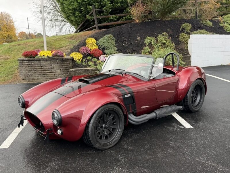 A red and black classic roadster convertible, reminiscent of classic muscle cars, is parked diagonally in a lot, with a landscaped embankment and colorful flowers in the background.