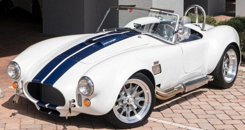 A white classic sports car with blue racing stripes is parked on a paved driveway. The car features chrome wheels, side pipes, and an open-top design—perfect for fans of high-performance cars.