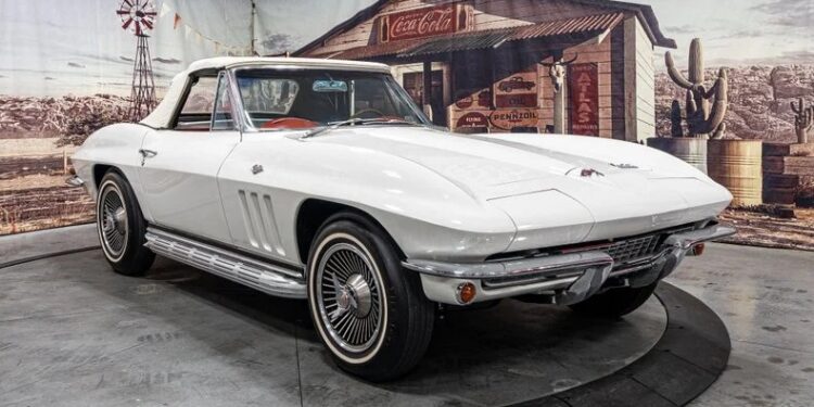 A classic white convertible car with a red interior is displayed indoors on a rotating platform, set against a vintage western-themed backdrop—perfect for enthusiasts of classic muscle cars.