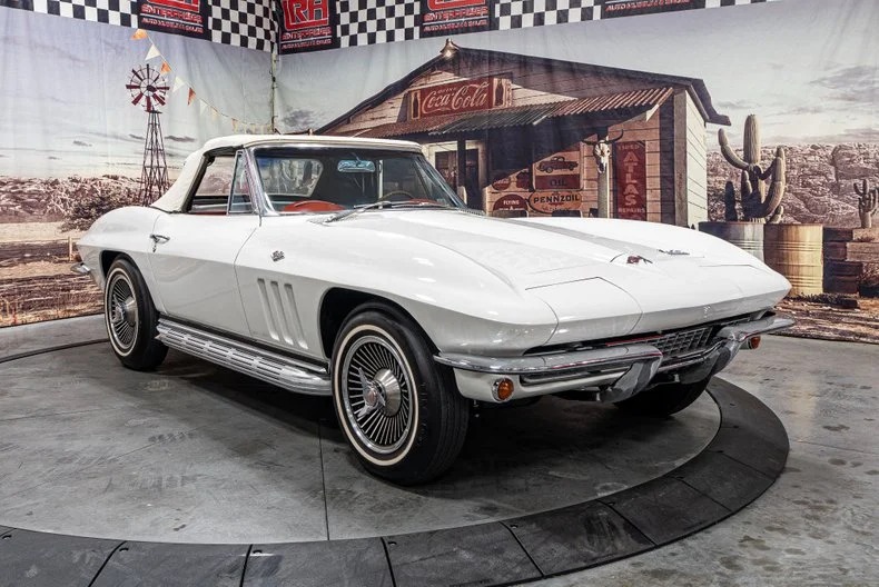 A classic white convertible car with a red interior is displayed indoors on a rotating platform, set against a vintage western-themed backdrop—perfect for enthusiasts of classic muscle cars.