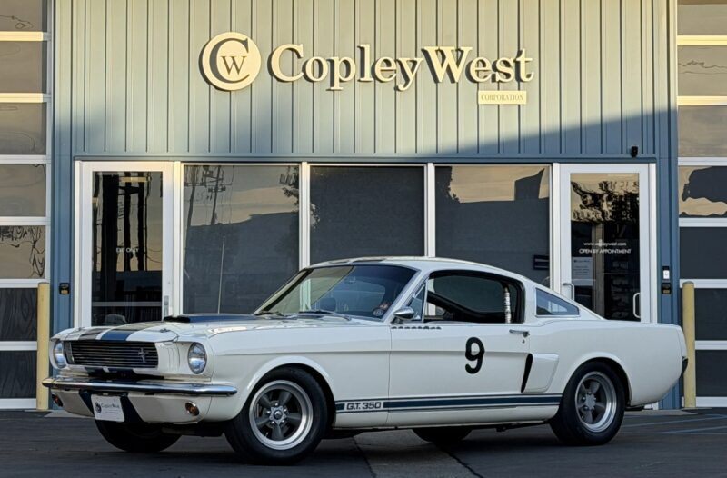A white classic Ford Mustang GT350 with racing stripes and the number 9 is parked in front of a building labeled “Copley West,” capturing the essence of high-performance cars and classic muscle cars.