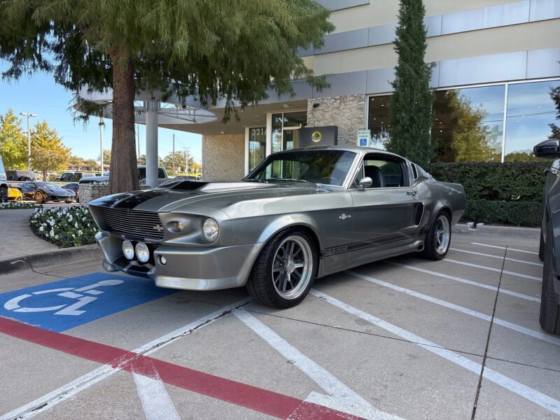 A silver classic muscle car Ford Mustang is parked in a handicapped space outside a modern building, partially over the marked access lines.