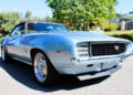 A light blue classic Chevrolet Camaro SS 427 convertible, a true icon among Classic Muscle Cars, is parked outdoors under bright sunlight with trees and greenery in the background.