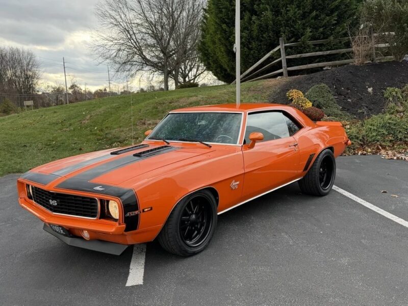 An orange 1969 Chevrolet Camaro SS with black racing stripes, a true icon among classic muscle cars, is parked in a lot near grass and trees.
