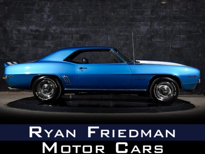 A blue classic Chevrolet Camaro coupe, a standout among Muscle Cars For Sale, is parked indoors on a black platform, with "Ryan Friedman Motor Cars" text displayed below the vehicle.
