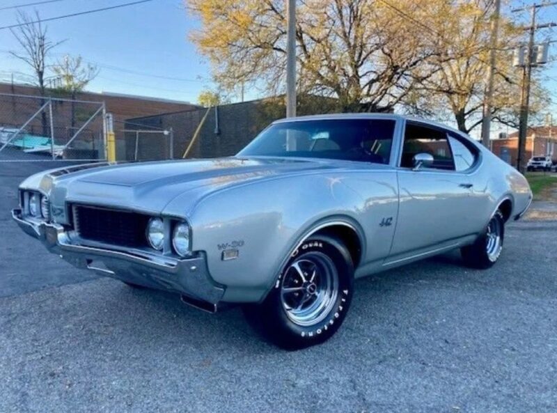 A silver classic muscle car, likely from the late 1960s or early 1970s, is parked on a paved lot near a fence and brick building with trees in the background—a striking example of high-performance cars from that era.