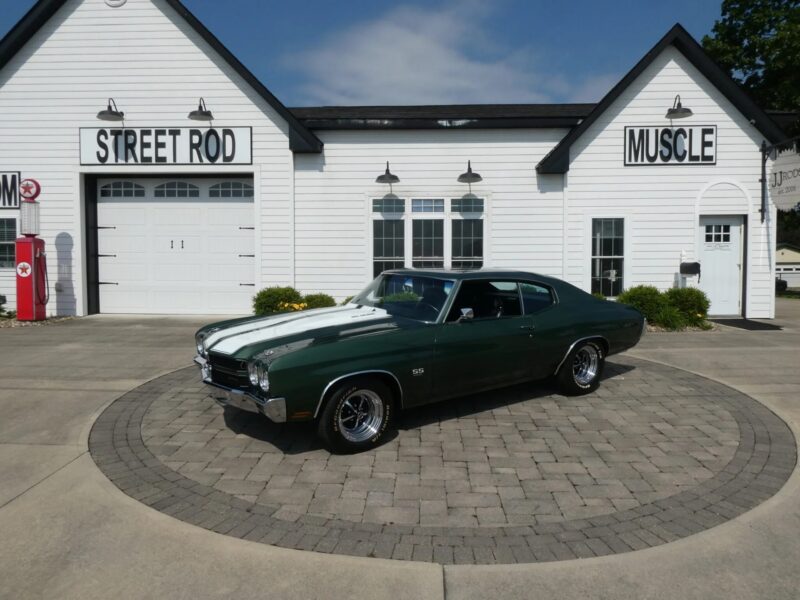A dark green classic muscle car with white racing stripes is parked in front of a white building labeled "STREET ROD" and "MUSCLE," showcasing the timeless appeal of high-performance cars.