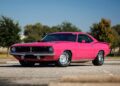 A bright pink classic muscle car, a true gem among Muscle Cars, parked on an empty lot with trees and buildings in the background under a clear blue sky.