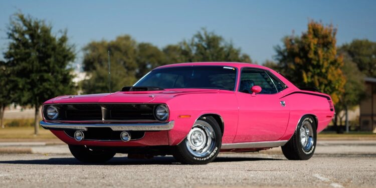A bright pink classic muscle car, a true gem among Muscle Cars, parked on an empty lot with trees and buildings in the background under a clear blue sky.