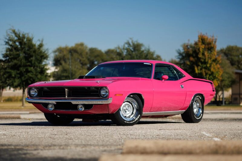 A bright pink classic muscle car, a true gem among Muscle Cars, parked on an empty lot with trees and buildings in the background under a clear blue sky.