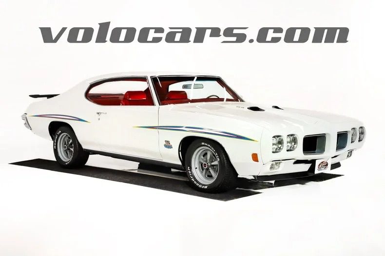 A white classic muscle car with red interior is displayed in a studio setting, with "volocars.com" written above the car, showcasing one of the finest Classic Muscle Cars for sale.
