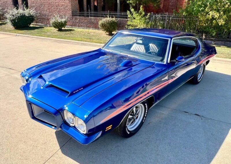 A blue classic muscle car, a true icon among High-Performance Cars, is parked on a sunlit street near a brick building and iron fence, viewed from a high front-side angle.