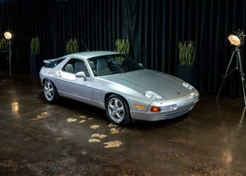 A silver Porsche 928 sports car, a true icon of 90s cars for sale, is parked indoors on a polished floor, with black curtains and potted plants in the background.