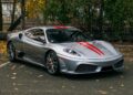 A silver Ferrari 430 Scuderia with red racing stripes, proudly displaying the prancing horse emblem, is parked in an outdoor lot surrounded by autumn trees and scattered leaves.