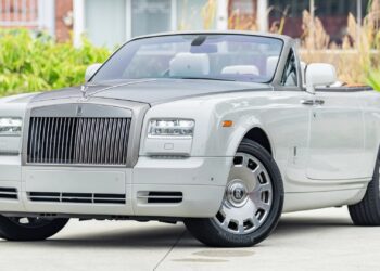 A silver Rolls-Royce convertible, a true luxury car, is parked outdoors on a paved surface with its roof down, surrounded by greenery and buildings in the background.