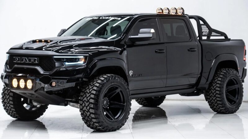 A black custom pickup truck with large off-road tires, lifted suspension, grille lights, and roof-mounted spotlights showcases its impressive capability in a bright indoor setting.