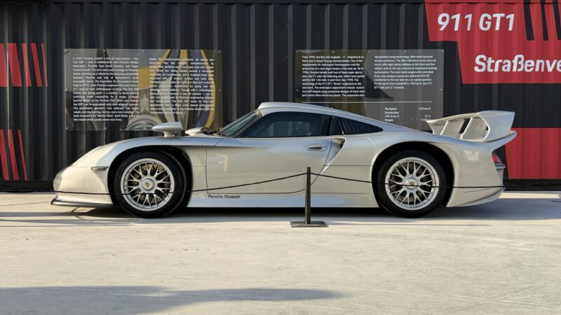 A silver Porsche 911 GT1 sports car is displayed outdoors, cordoned off with black rope in front of a black and red shipping container with informational boards. Feature Exclusive: 2025 Icons of Porsche Dubai.