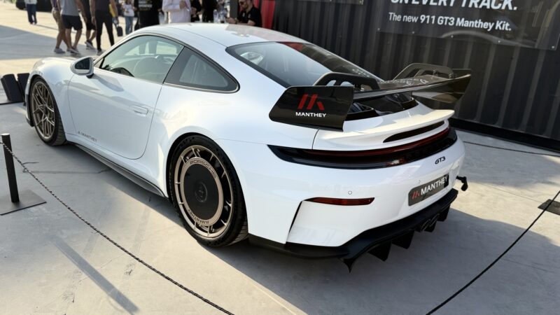 Feature Exclusive: A white Porsche 911 GT3 with a Manthey kit is displayed outdoors at the 2025 Icons of Porsche Dubai, featuring a large rear wing and distinctive wheels.