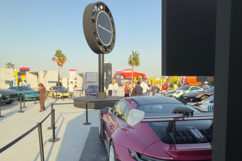 Feature Exclusive: 2025 Icons of Porsche Dubai—several sports cars are displayed outdoors at an event, with people walking around, palm trees, and branded signage visible in the background.