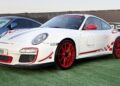 Feature Exclusive: This white Porsche sports car with red accents and detailing, including striking red wheels and side decals, is showcased on green turf at the 2025 Icons of Porsche Dubai outdoor event.