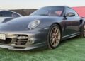 A grey Porsche sports car with gold rims, featured in the Exclusive: 2025 Icons of Porsche Dubai, is parked on green artificial grass near a white car, set against a striking pink and black background.