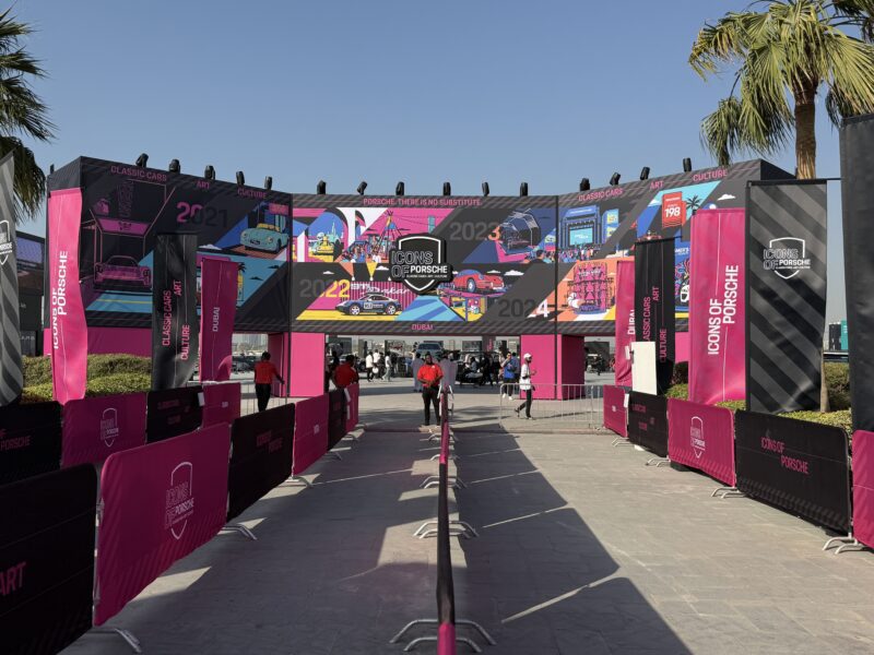 Entrance to an outdoor event with "Icons of Porsche" banners, colorful graphics, and people walking along a fenced pathway under sunny weather—Feature Exclusive: 2025 Icons of Porsche Dubai.