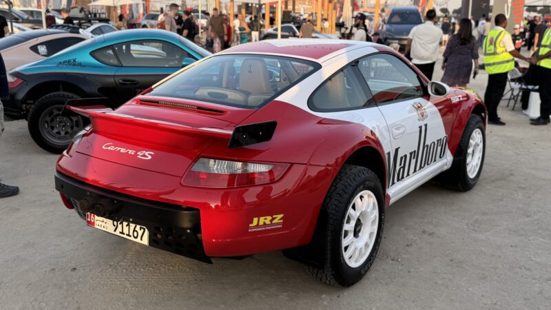 Feature Exclusive: This red and white Porsche Carrera 4S rally car with Marlboro livery is parked outdoors at an event, as part of the 2025 Icons of Porsche Dubai, with people and other vehicles in the background.