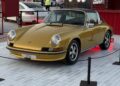 Feature Exclusive: 2025 Icons of Porsche Dubai—a classic gold Porsche sports car is displayed on a tiled platform, roped off from the public, with other vehicles and a red structure in the background.