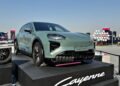 A light green Porsche Cayenne Turbo is displayed on a platform outdoors, featured as part of the 2025 Icons of Porsche Dubai event, with signage and other displays visible in the background.