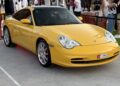 Feature Exclusive: A yellow Porsche 911 sports car is parked on a concrete surface, featured at the 2025 Icons of Porsche Dubai event, with people standing in the background.