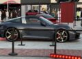 A dark gray Porsche 911 Targa is displayed outdoors at a car show, surrounded by barriers and parked on a tiled surface with other cars and people in the background. Feature Exclusive: 2025 Icons of Porsche Dubai.